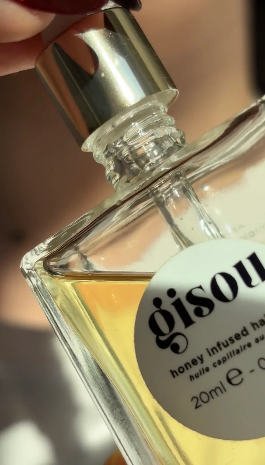 Close-up of a partially opened Gisou hair oil bottle with Angelina softly blurred in the background